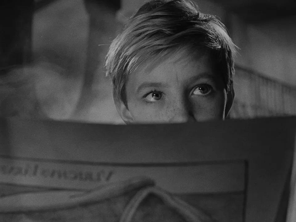 Ivan’s Childhood (1962) movie still: closeup — A child's face peeks over the edge of a large open newspaper. The individual holds the paper with…