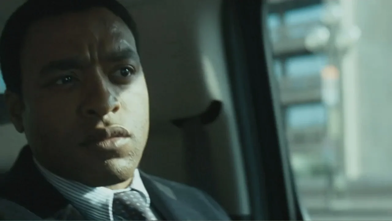 Salt (2010) movie still: closeup — A man sits inside a vehicle while passing through an urban environment during daylight. He wears a dark suit and…