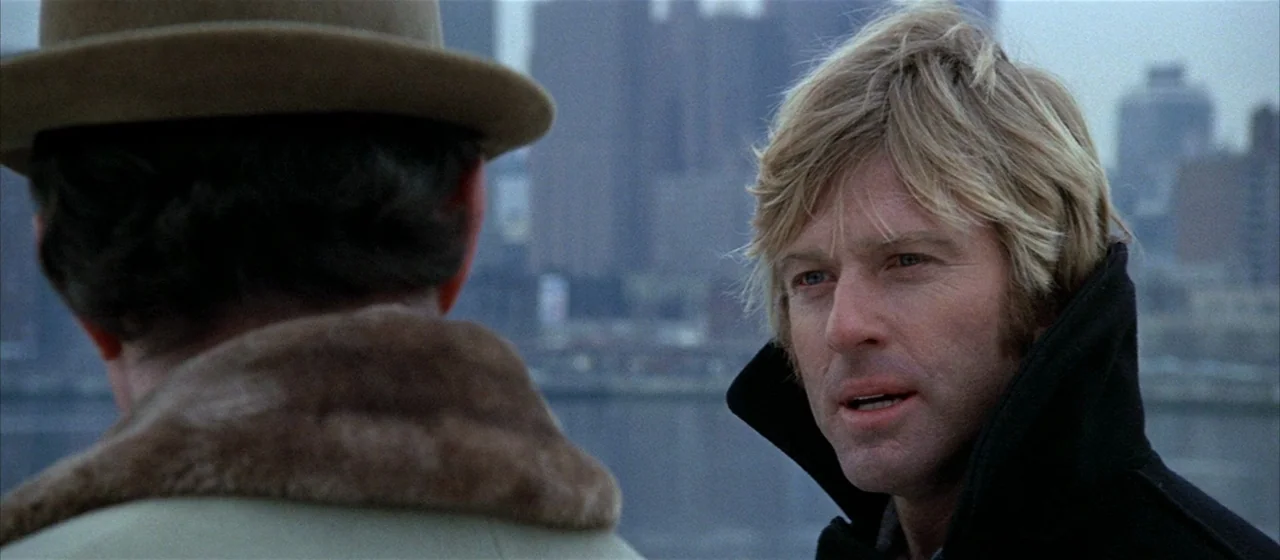 Three Days of the Condor (1975) movie still: medium — Two men stand near the water with a blurred city skyline in the distance. The man on the right…