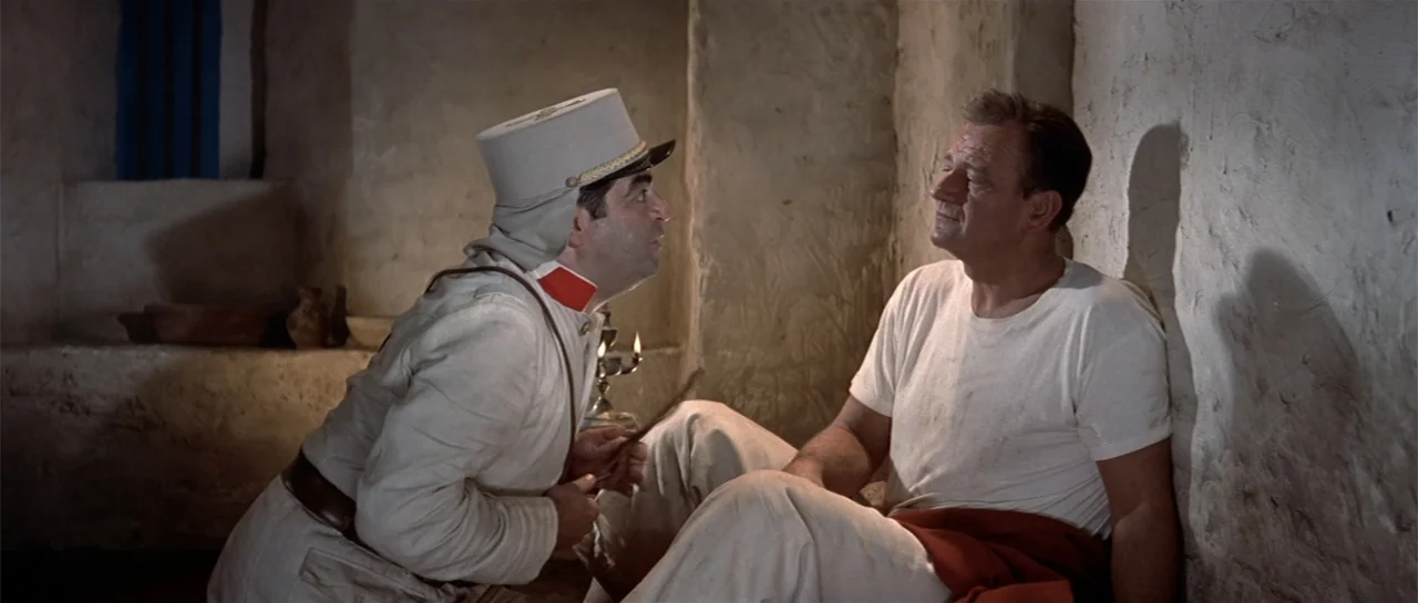 Legend of the Lost (1957) movie still: medium — A rustic stone room features textured walls and minimal furnishings. Two men in historical clothing…
