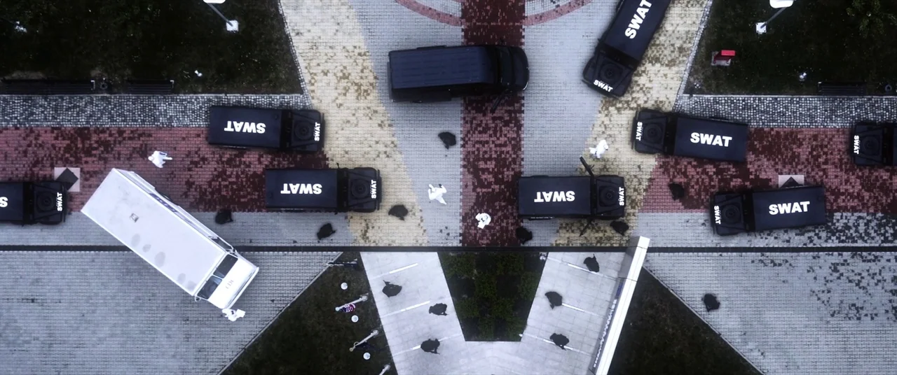 Mayhem (2017) movie still: establishing — A top-down view shows multiple black SWAT vehicles surrounding a central paved area. Several individuals…