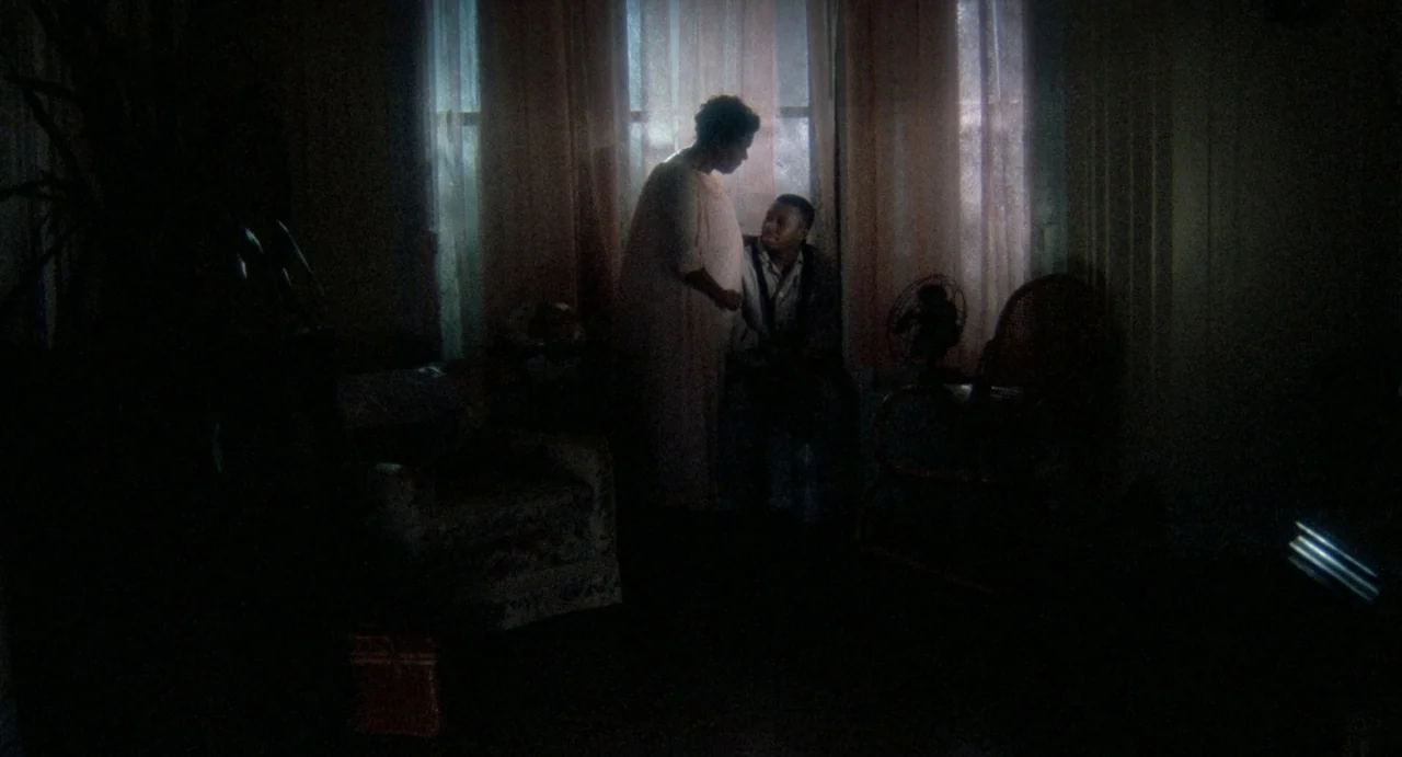 Def By Temptation (1990) movie still: medium — A dimly lit room with draped windows and dark furniture silhouetting the subjects. A man sits in a…
