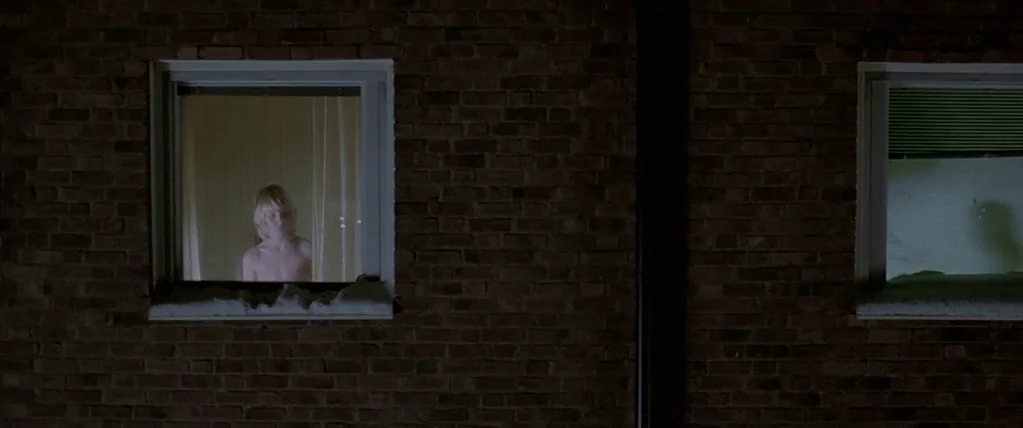 Let The Right One In (2008) movie still: wide — A dark brick building wall at night features two recessed square windows. A shirtless young boy…