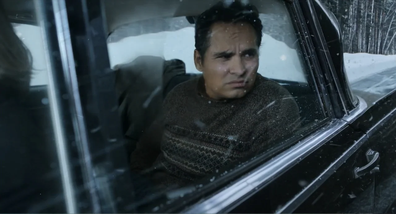 A Million Miles Away (2023) movie still: medium — A man sits inside a dark sedan traveling past a snowy forest landscape. He looks out the side…
