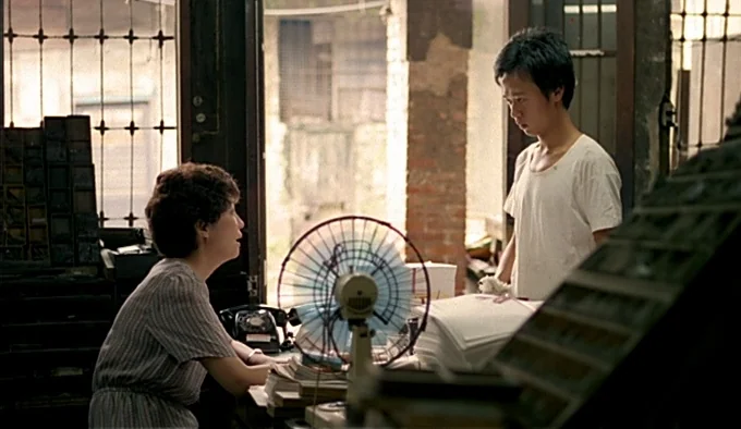 Dust In The Wind (1986) movie still: medium — A cluttered workspace filled with stacked paper, metal shelving, and a small desk fan. A woman sits…