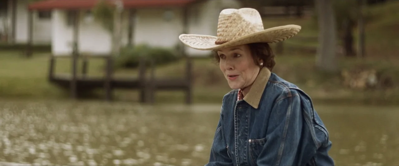Reagan (2024) movie still: medium — A woman stands outdoors in front of a calm body of water. She wears a straw hat and a denim jacket while looking…