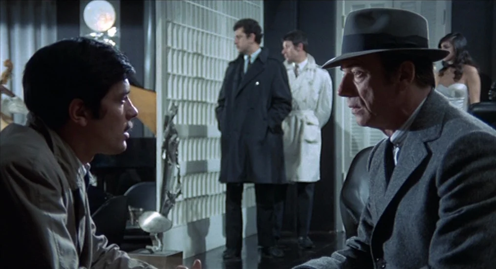 Le Cercle Rouge (1970) movie still: medium — A modern lounge with dark walls and a large, textured white architectural divider screen. Two men face…