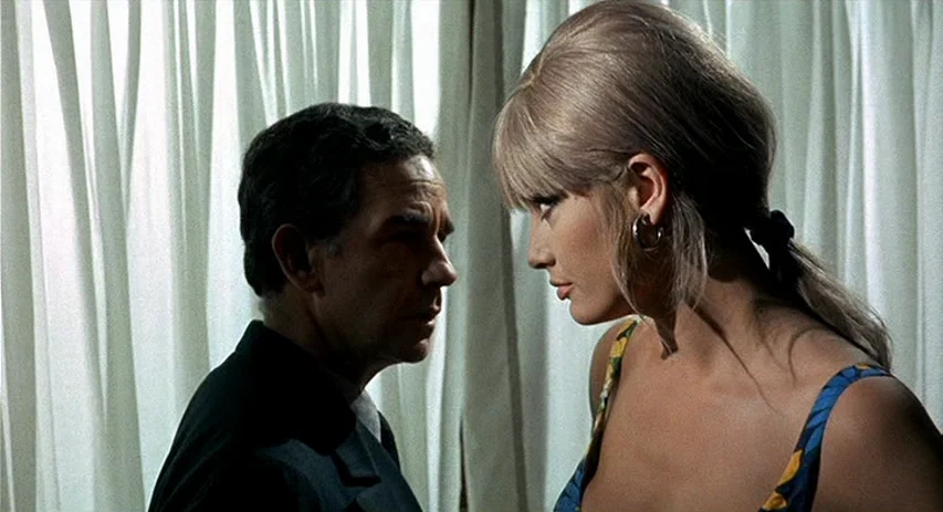 Perversion Story (1969) movie still: medium — A man and a woman stand inside near a large white curtained window. The woman with blonde hair and the…