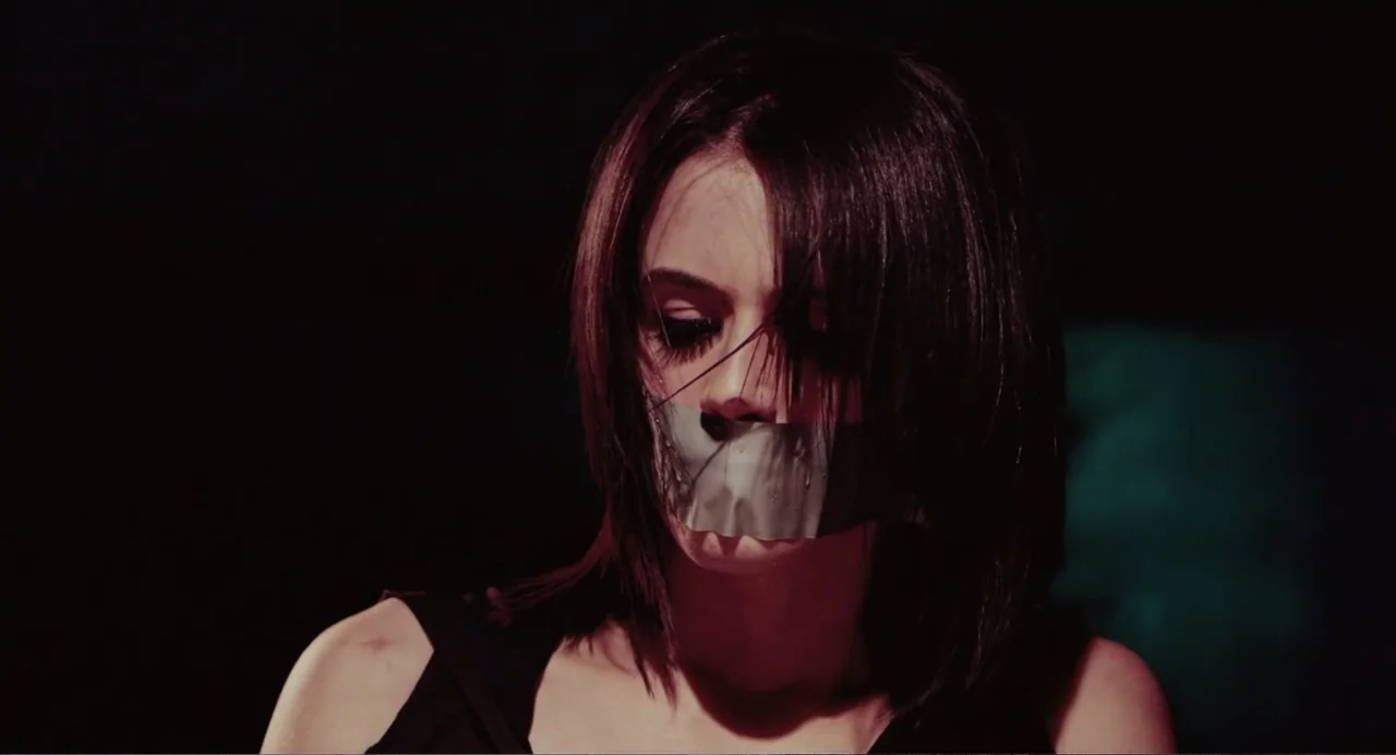 Terrifier (2018) movie still: closeup — A dark, nondescript interior space with deep shadows surrounding a central subject. A young woman with dark…