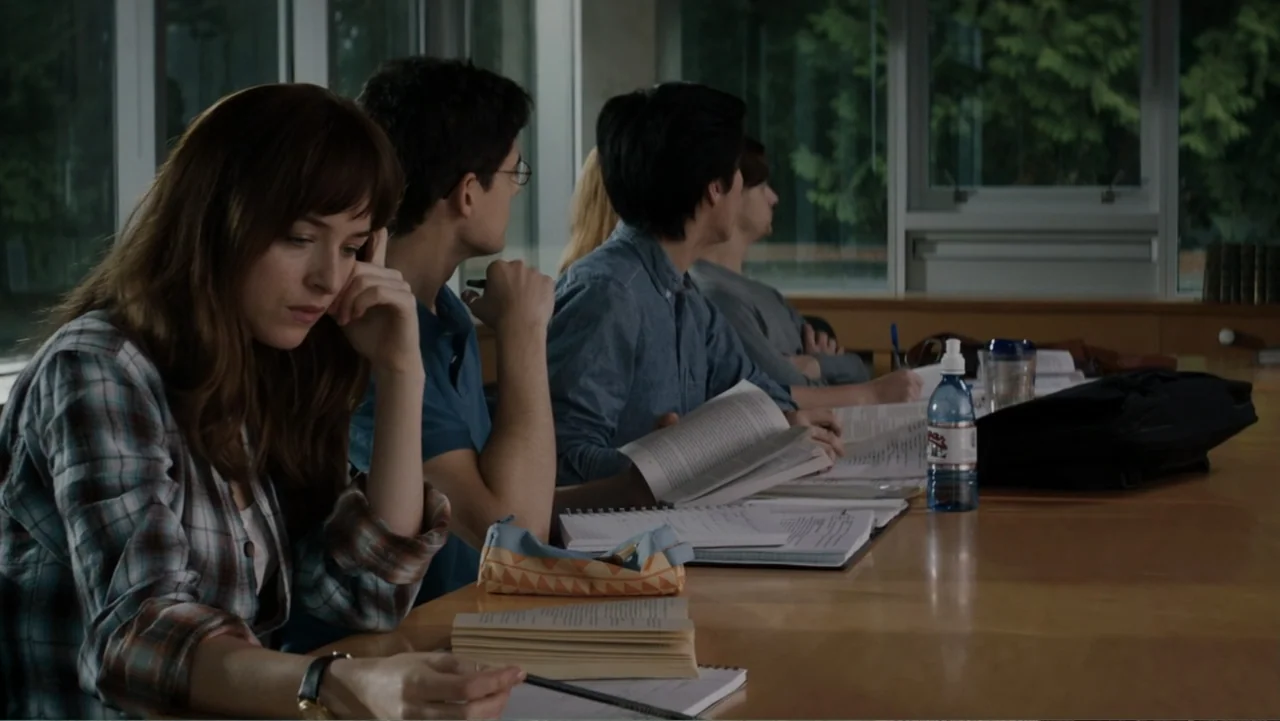 Fifty Shades of Grey (2015) movie still: medium — A student sits at a wooden table in a brightly lit library setting. A woman in a flannel shirt…