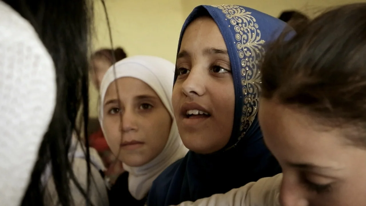 City of Two Springs (2019) movie still: medium — A group of young girls interact inside a room with plain light-colored walls. They are gathered…
