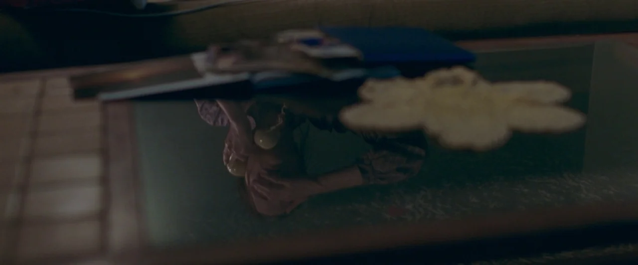 Censor (2021) movie still: closeup — A glass-topped table holds scattered photographs and a decorative doily. A distorted reflection of a person…