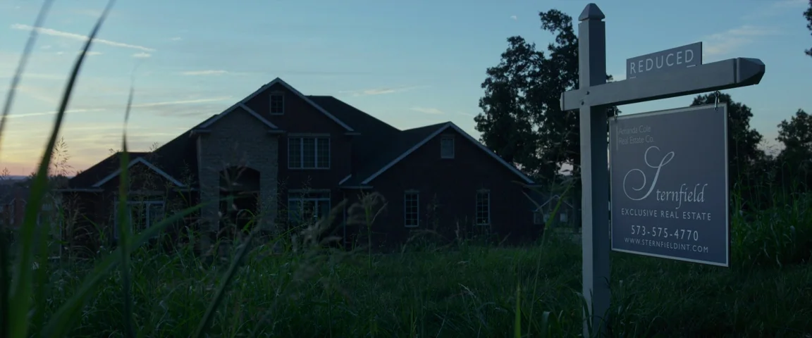 Gone Girl (2014) movie still: wide — A dark residential house stands in the background at dusk under a blue sky. A real estate sign with the name…