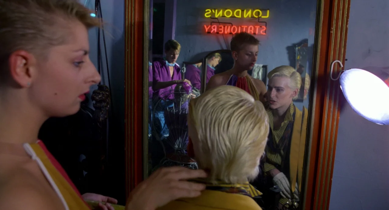 Liquid Sky (1982) movie still: medium — A dimly lit room features a large mirror framed in decorative wood and gold. A person stands behind another…