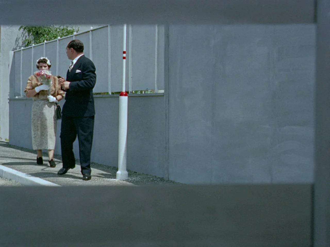 Mon Oncle (1958) movie still: wide — A man and a woman walk along a concrete sidewalk beside a tall fence. The scene is framed by dark horizontal…