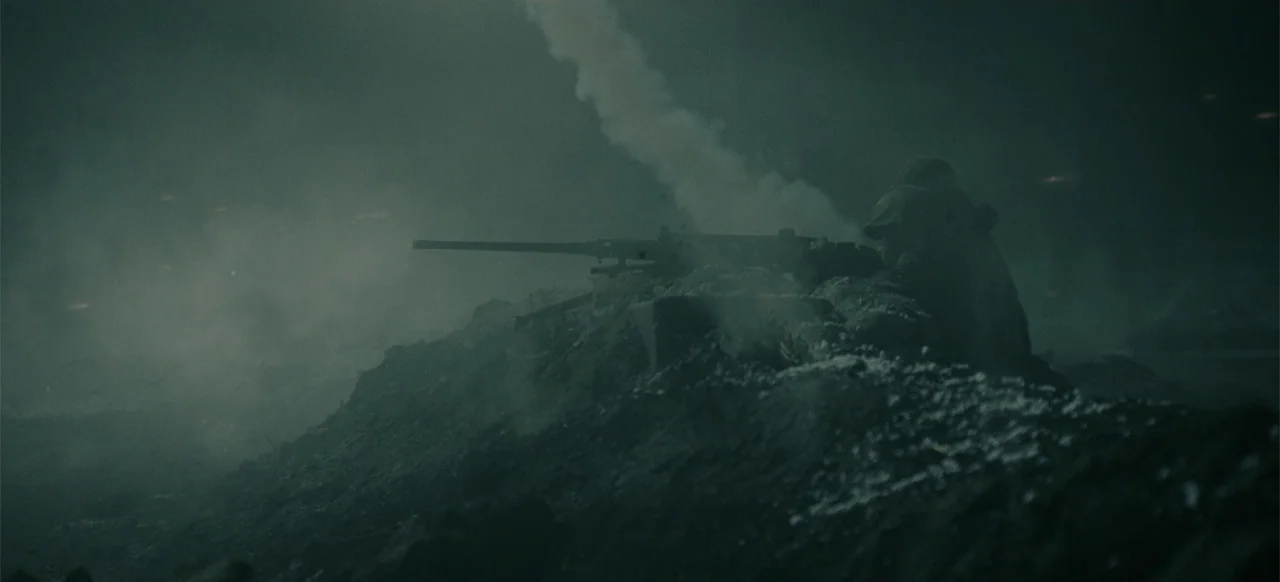 Devotion (2022) movie still: wide — A dark, smoke-filled battlefield features a soldier operating a machine gun on an embankment. Thick plumes of…