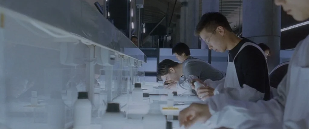Code 46 movie still: medium — Technicians in white lab coats work at a sterile table illuminated by bright light. Several workers stand side-by-side…