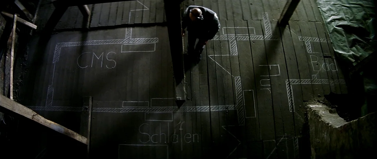 The Lives of Others (2006) movie still: wide — A dimly lit room with wooden floorboards marked with white chalk architectural floor plans. A man…