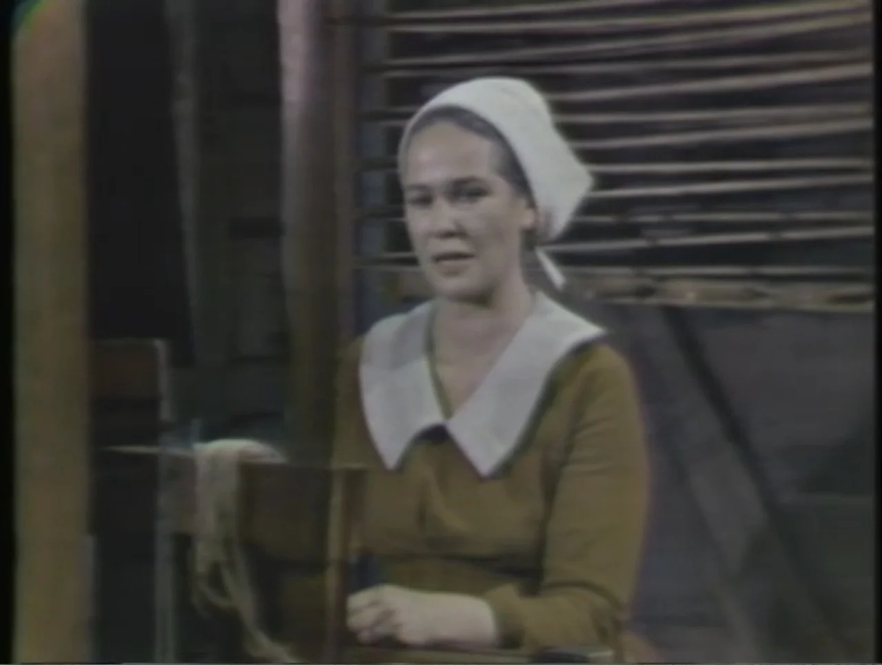 The Crucible (1996) movie still: medium — A dimly lit interior space with a wooden loom structure in the background. A woman in period clothing sits…