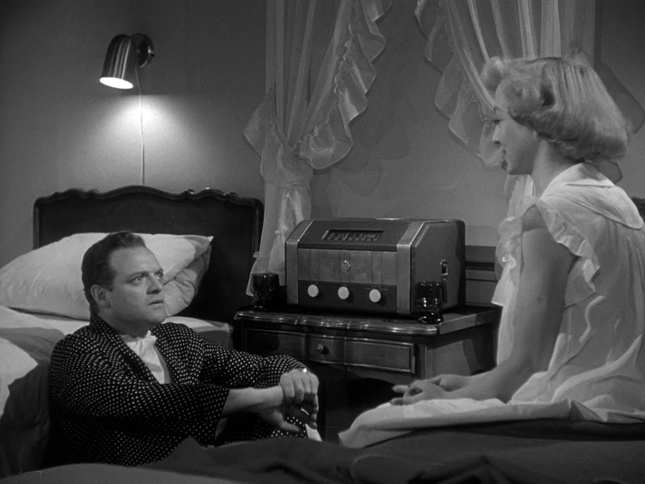 The Prowler (1951) movie still: medium — A 1950s bedroom with a dark wood headboard and sheer white ruffled curtains. A man in a polka-dot robe sits…