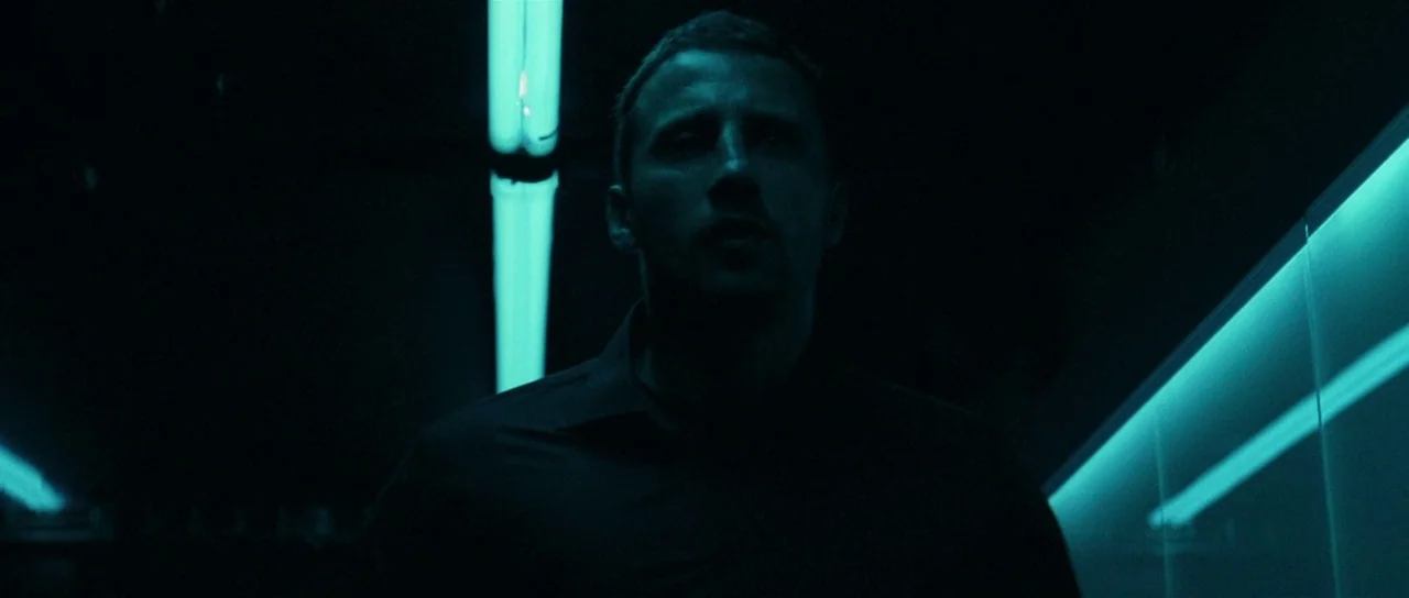 Bullhead (2011) movie still: medium — A dark, narrow corridor is illuminated by harsh cyan fluorescent light fixtures on the ceiling. A man walks…