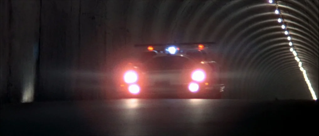 THX 1138 (1971) movie still: wide — A dark concrete tunnel has a single line of bright lights on one side. A silver sports car drives directly…