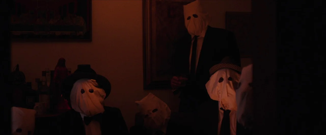 Fréwaka (2024) movie still: medium — A dark, dimly lit room features framed artwork on the walls and a side table. Several figures wearing formal…
