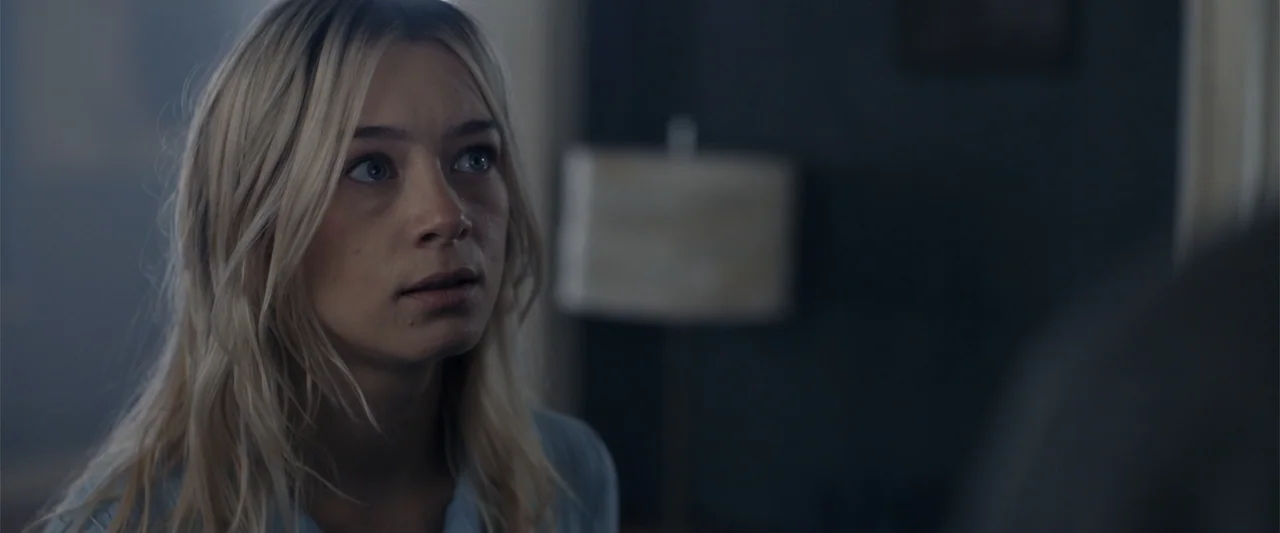 The Good Neighbor (2022) movie still: closeup — A dimly lit room contains a person seen over the shoulder of another. The young woman looks intently…