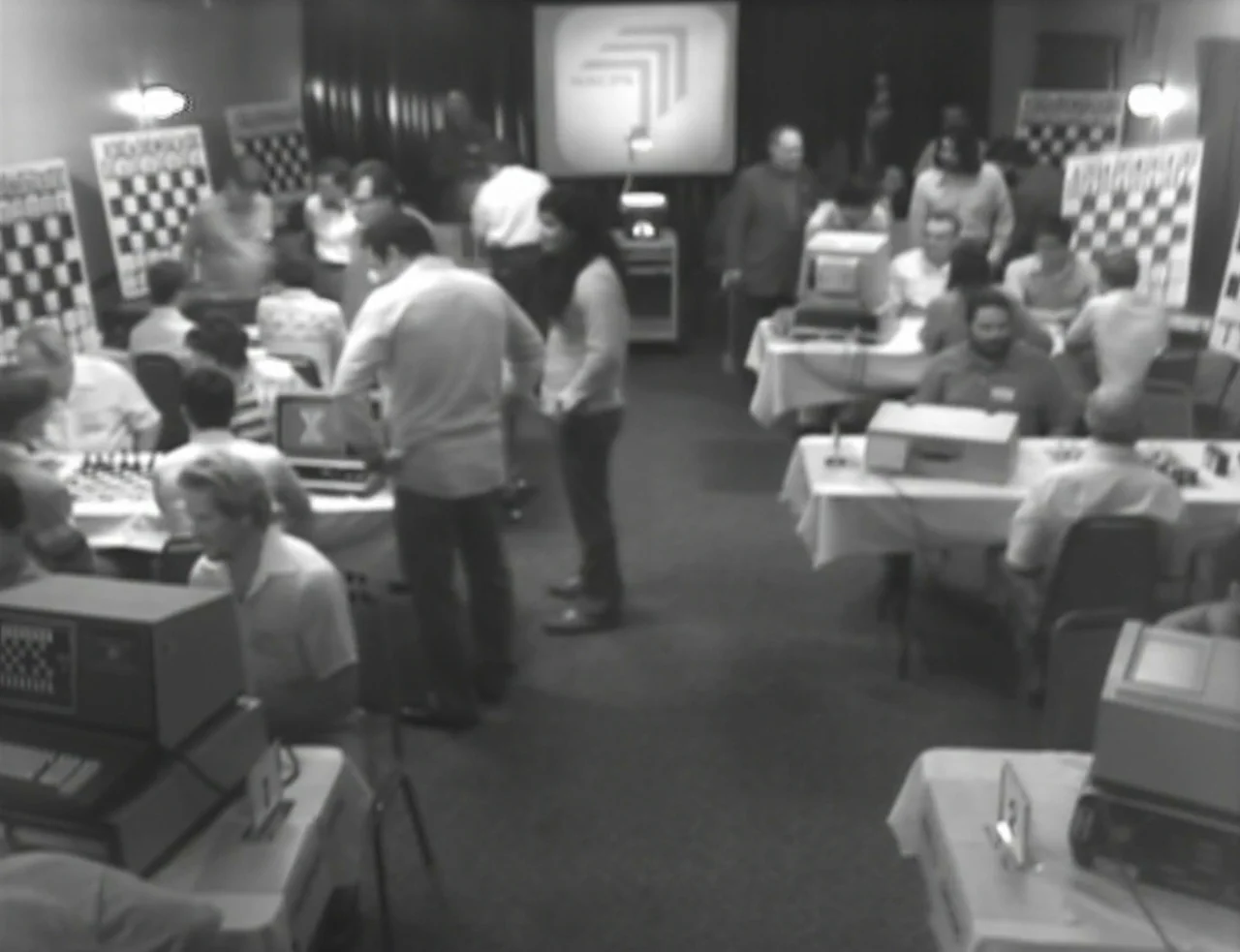 Computer Chess (2013) movie still: wide — A crowded room is filled with rows of tables featuring vintage computing equipment and chessboards. People…