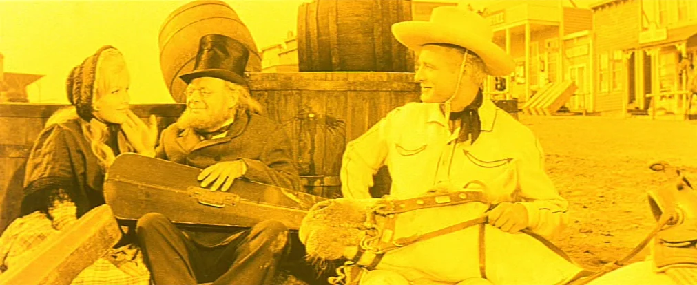 Lemonade Joe (1964) movie still: medium — A group of three people sit near wooden barrels in an old western town. The man in the cowboy hat holds a…