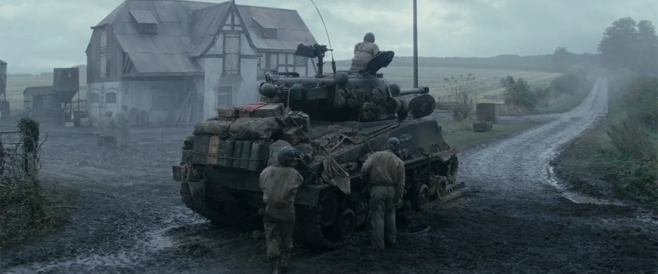 Fury (2014) movie still: wide — A mud-covered dirt road stretches past a timber-framed house toward an open field. Three soldiers gather around a…
