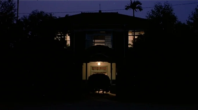 A Summer at Grandpa’s (1984) movie still: establishing — A two-story dark residential house is surrounded by silhouettes of dense trees at night. A…