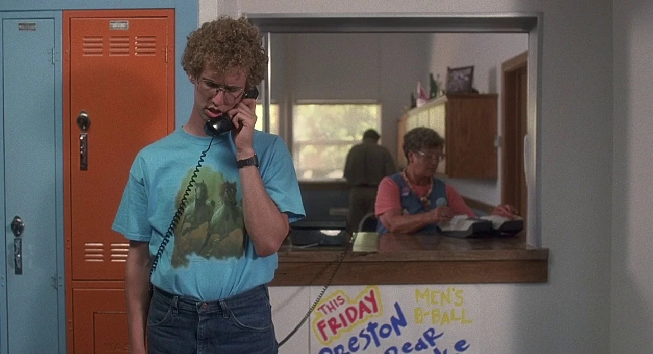 Napoleon Dynamite (2004) movie still: medium — A school hallway features orange lockers and a wall with handwritten signs. A young man stands in the…