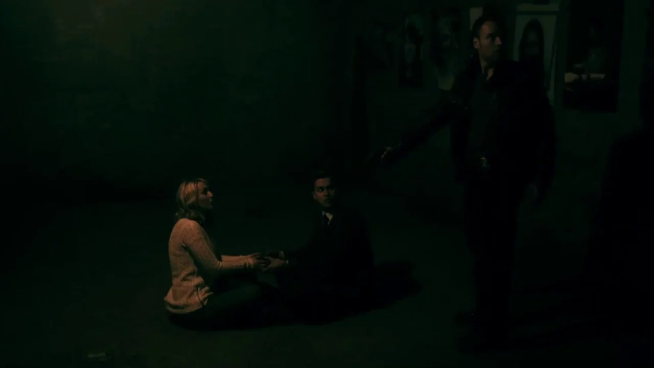 Hellraiser: Judgement (2018) movie still: wide — A dimly lit room with dark walls and posters on the far wall. A man holding a handgun stands over a…