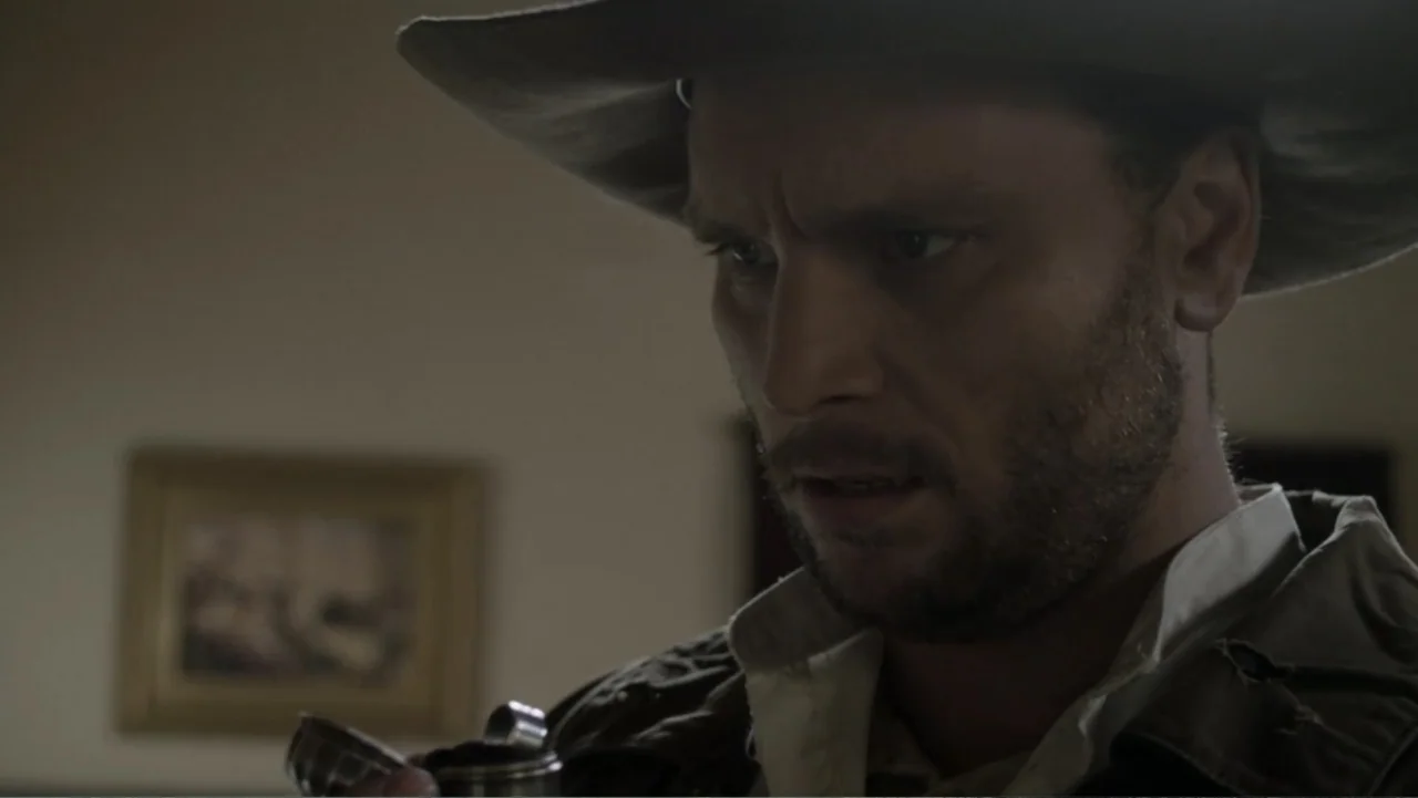 Dawn Rider (2012) movie still: closeup — A dimly lit room with an out-of-focus painting hanging on a plain wall. A man in a dusty cowboy hat looks…