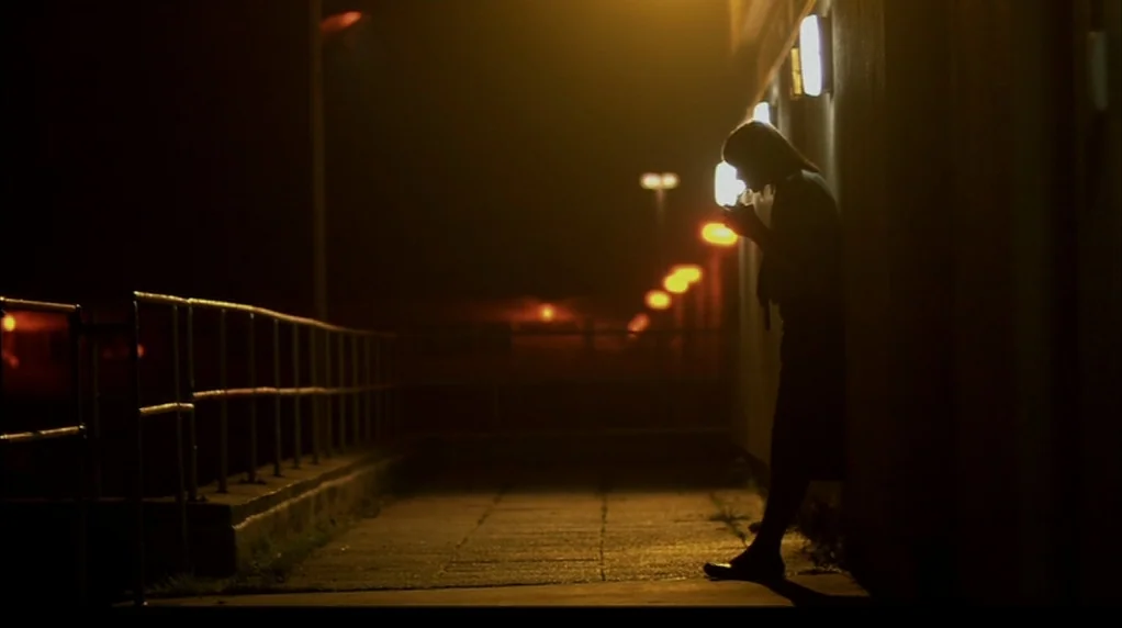 Red Road (2006) movie still: medium — A concrete walkway at night illuminated by a series of glowing yellow streetlights. A solitary figure stands…