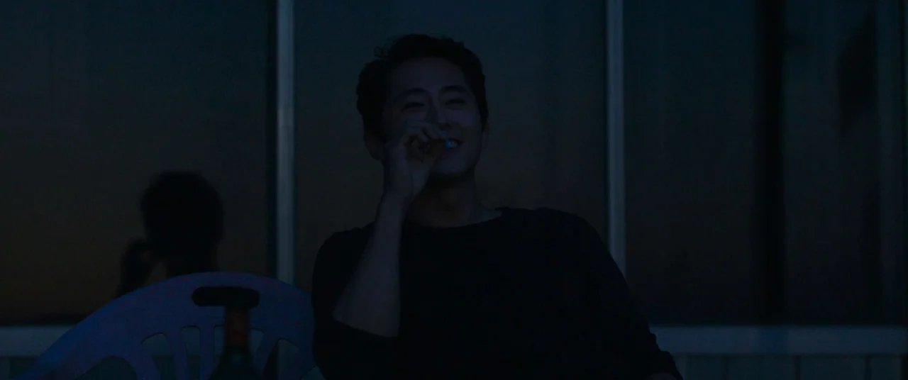 Burning (2018) movie still: medium — An outdoor patio at night features a man seated before a glass sliding door. He holds a small item toward his…