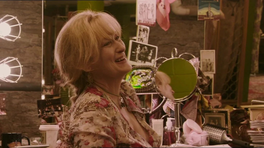 A Prairie Home Companion movie still: medium — A cluttered dressing room vanity is covered in personal photographs and various cosmetic items. A…