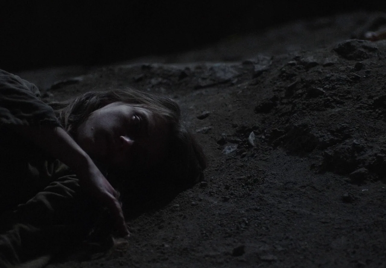 You Won’t Be Alone (2022) movie still: closeup — A person lies on a dark patch of uneven, rocky dirt ground. The subject rests their head on their…