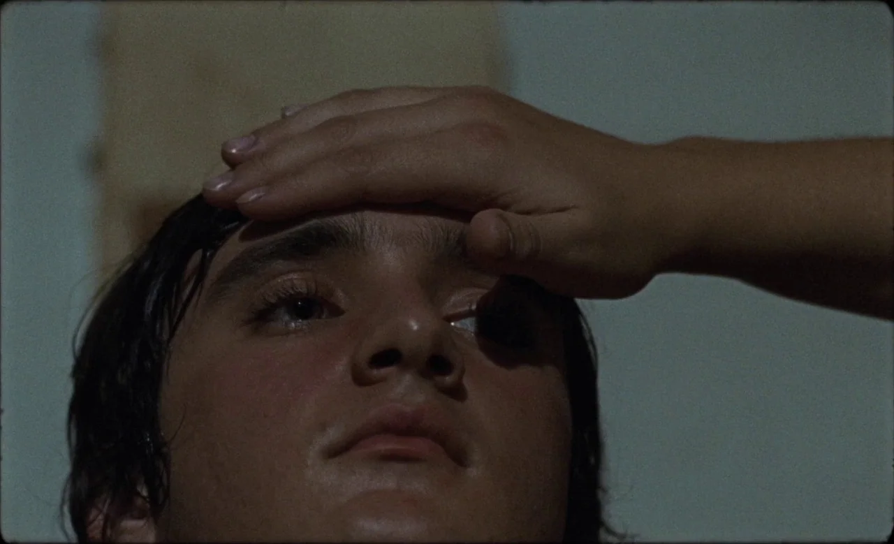 Happy as Lazzaro (2018) movie still: closeup — A close-up of a young male face set against a plain wall. A hand reaches into the frame to rest on…