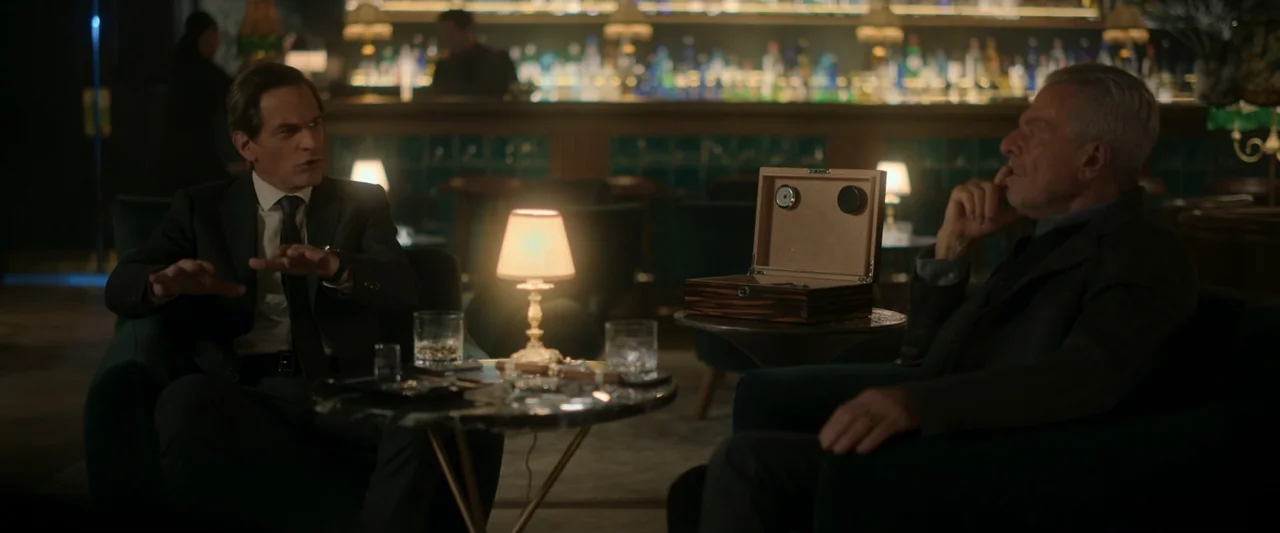 Doublespeak (2021) movie still: medium — A dimly lit upscale bar lounge features plush seating and a distant illuminated back bar. Two men in dark…
