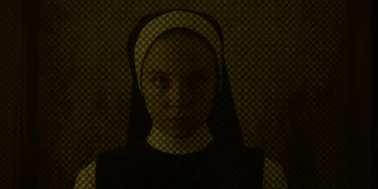 Immaculate (2024) movie still: closeup — A dark confessional booth features a fine metal screen between the camera and subject. The nun wearing a…