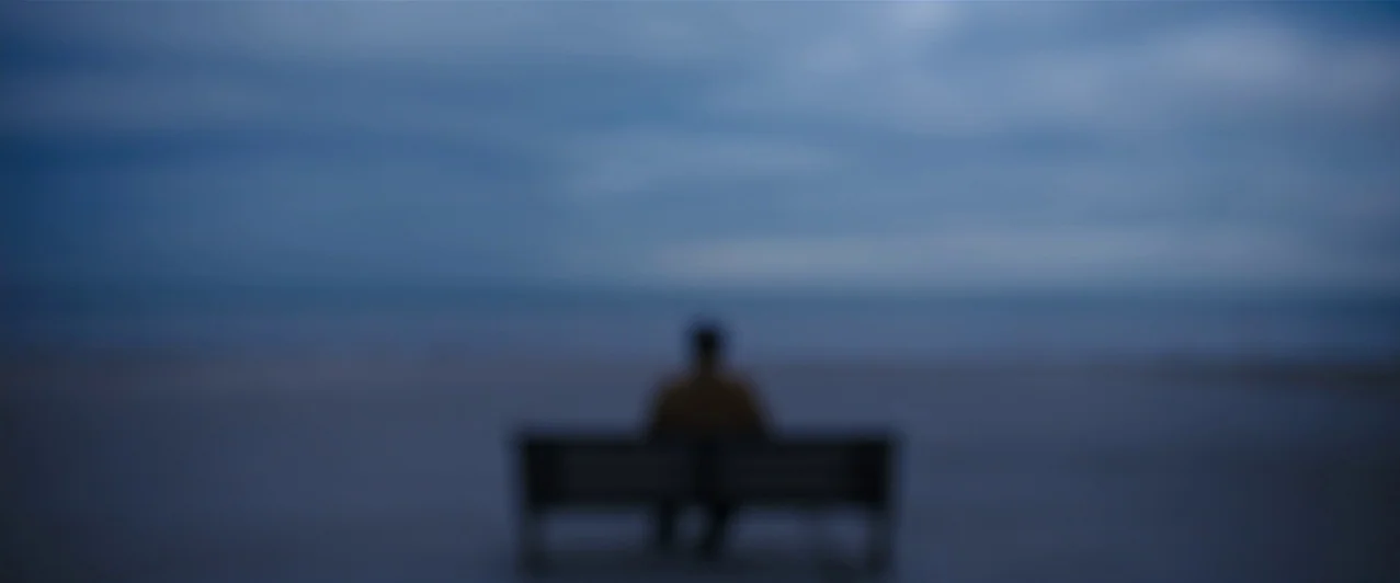 Ghost Stories (2017) movie still: wide — A beach at blue hour features a single park bench positioned on the sand. A lone figure sits on the bench…