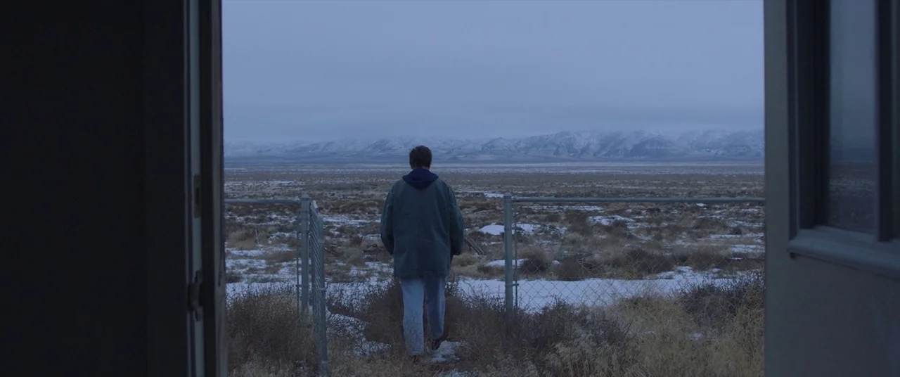 Nomadland (2020) movie still: wide — A man walks away from a building entrance into a vast, snowy desert landscape. In the distance, low mountains…