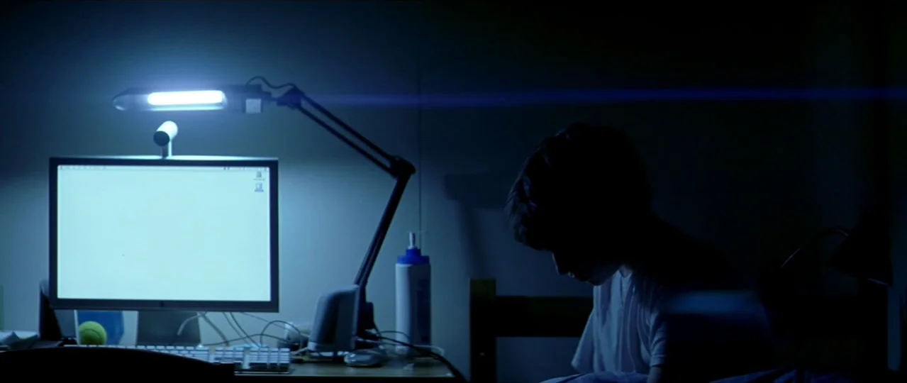 Afterschool (2008) movie still: medium — A dark, dimly lit room contains a computer setup with a bright monitor screen. A young male sits in…