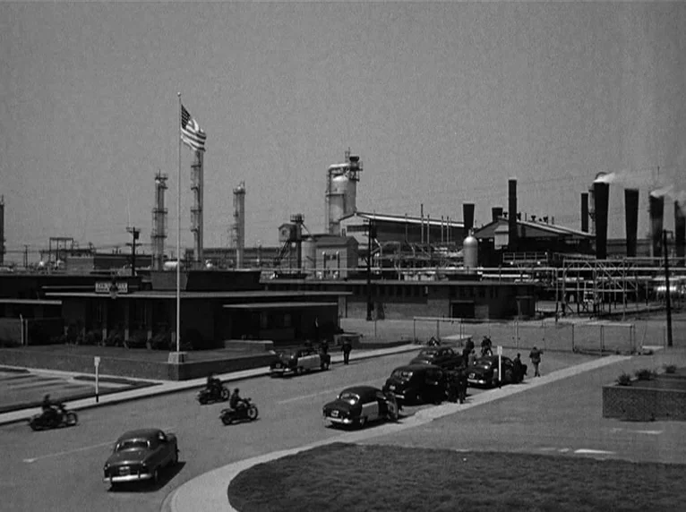 White Heat (1949) movie still: establishing — A sprawling industrial plant with tall smokestacks and piping sits behind a paved entrance. Several…