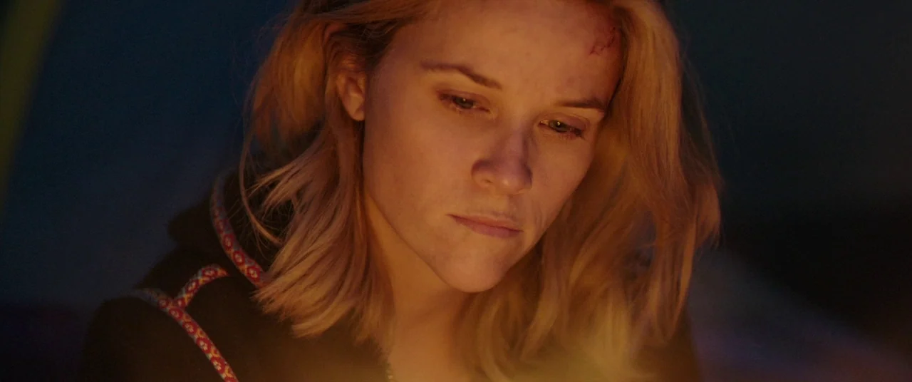 Wild (2014) movie still: closeup — A dark, outdoor setting is dimly lit by a nearby fire at night. A woman gazes downwards at the light while…