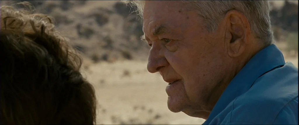 Into the Wild (2007) movie still: closeup — An elderly man in a blue shirt stands in an expansive, dry desert landscape. He is turned in side…