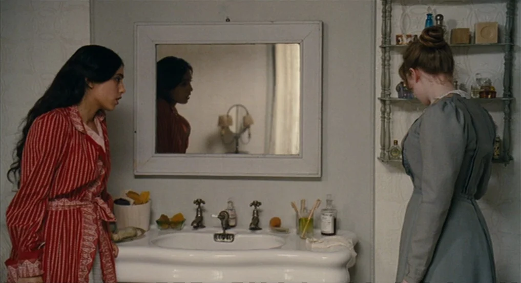 House of Tolerance (2011) movie still: medium — A 19th-century bathroom features a porcelain pedestal sink beneath a framed wall mirror. Two women…