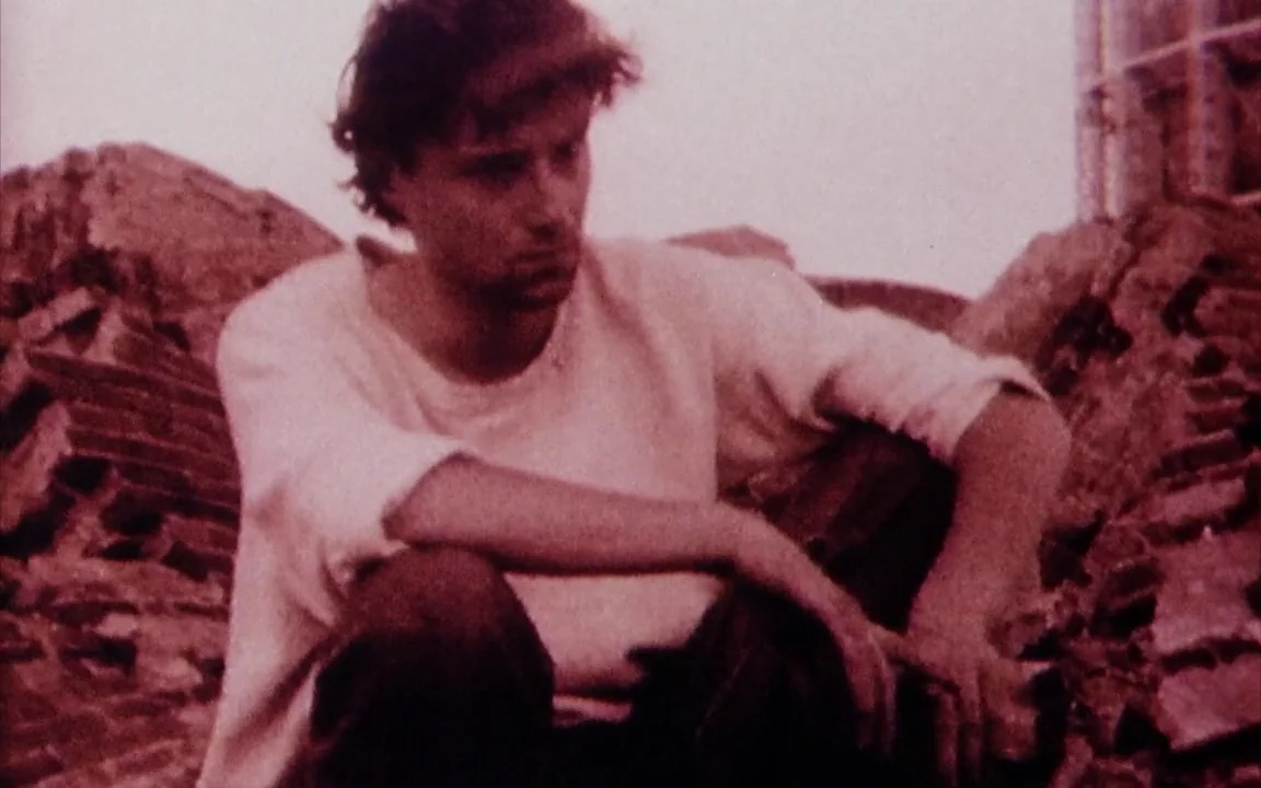 The Last of England (1987) movie still: medium — A pile of discarded red bricks and rubble forms the setting for the subject. A young man sits among…